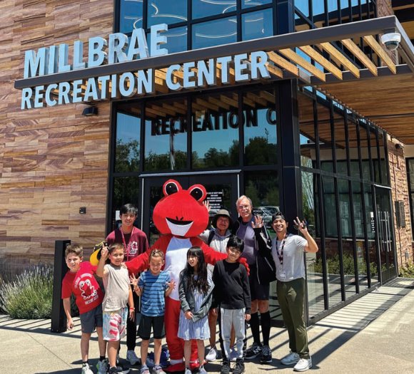 Picture of community members of all ages waving in front of Millbrae Recreation Center with Millie. 